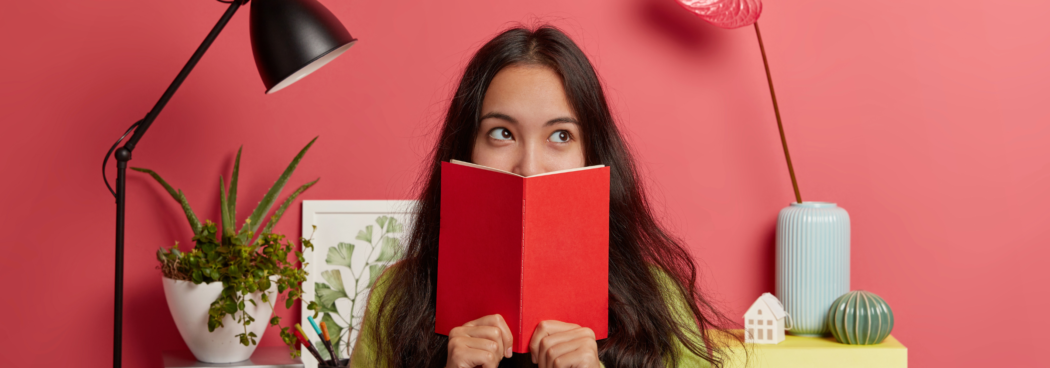 Girl reading a book with a thoughtful expression, sitting at a desk with study materials and plants around her.