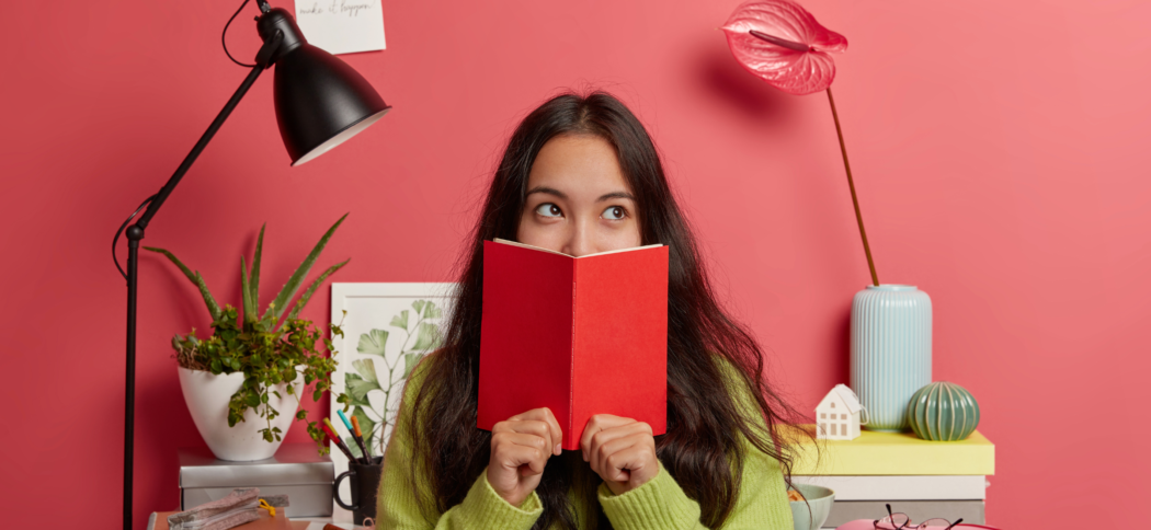 Girl reading a book with a thoughtful expression, sitting at a desk with study materials and plants around her.