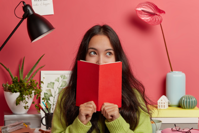 Girl reading a book with a thoughtful expression, sitting at a desk with study materials and plants around her.